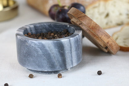 Kleine runde Gewürzschale aus dunkelgrau-weißem Marmor mit Holzdeckel, präsentiert auf einem elegant gedeckten Tisch.